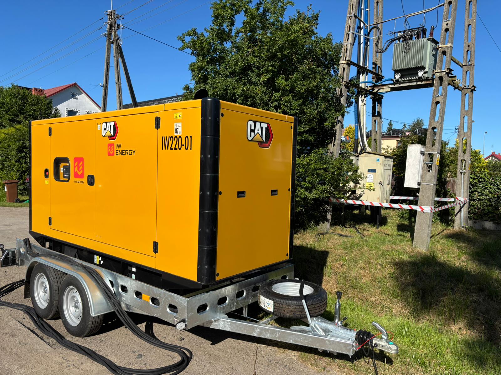 Żółty agregat prądotwórczy CAT IW220-01 od IW Energy, zamontowany na dwuosiowej przyczepie, podłączony do infrastruktury elektrycznej przy słupie z transformatorem. Ustawiony na ulicy w pobliżu budynków mieszkalnych i zieleni.
