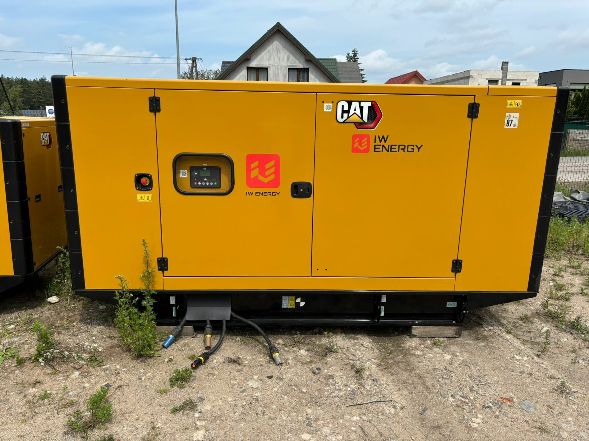 Stacjonarny agregat prądotwórczy CAT wynajmowany przez IW Energy, ustawiony na placu z widocznymi kablami podłączeniowymi, w tle zabudowa jednorodzinna.