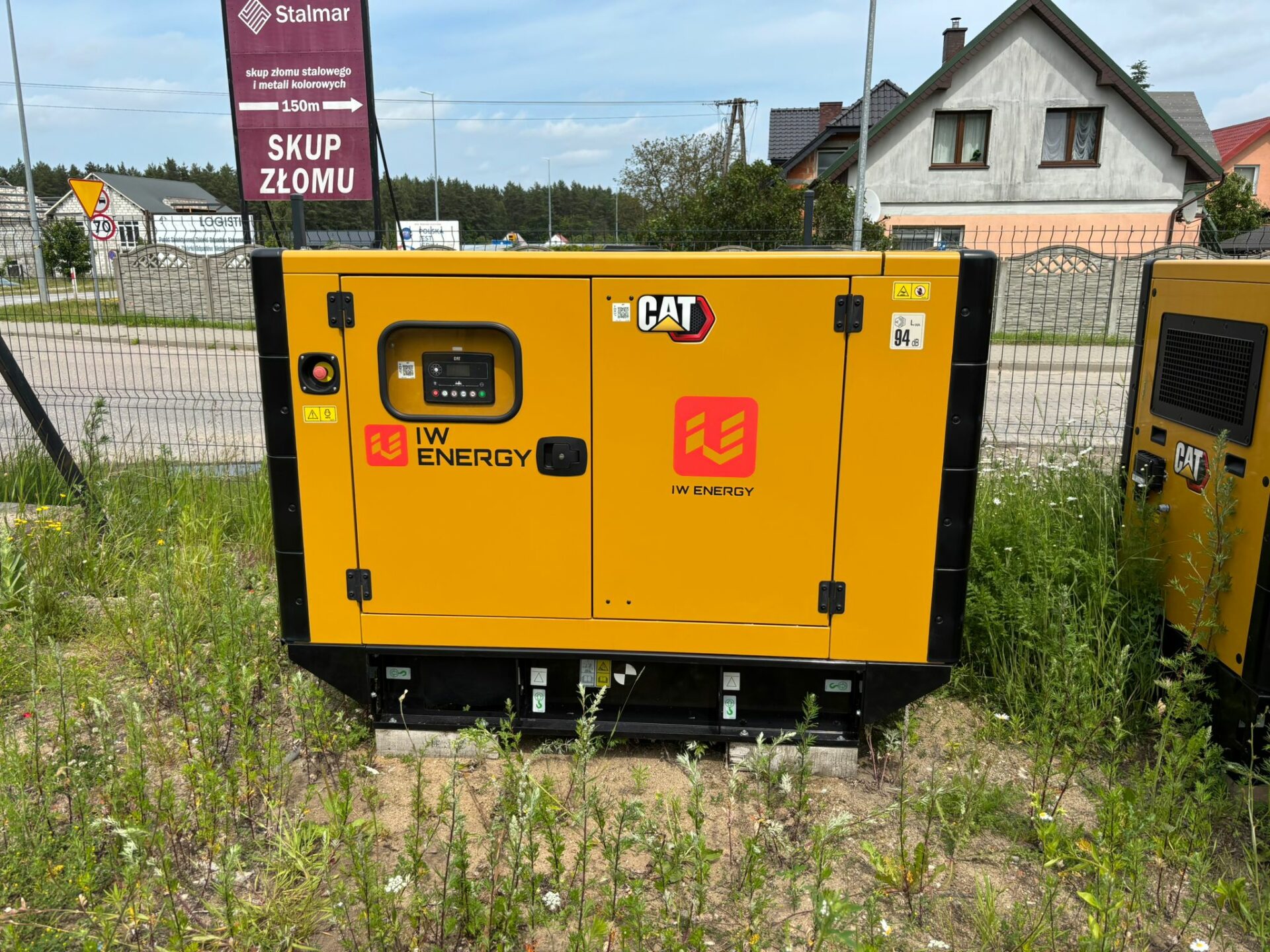 Agregat prądotwórczy CAT wynajmowany przez IW Energy, ustawiony na betonowych podporach, widoczny w otoczeniu ogrodzenia i zabudowy jednorodzinnej. Urządzenie oznaczone logotypami CAT i IW Energy.